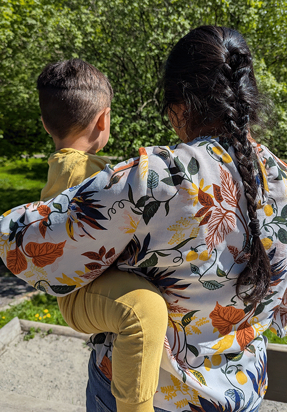 En kvinne med lang flette, iført en blomstermønstret bluse, bærer et lite barn kledd i gult mens hun går utendørs på en solskinnsdag. Frodige, grønne trær og sollys er synlige i bakgrunnen.