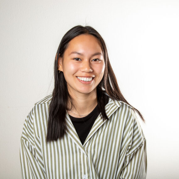 En ung kvinne med langt, rett, mørkt hår smiler til kameraet. Hun har på seg en grønn og hvit stripete skjorte over en svart topp, og poserer mot en ensfarget, lys bakgrunn.