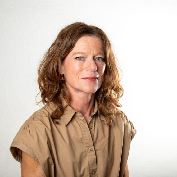 En kvinne med bølget, skulderlangt brunt hår og lys hud har på seg en brun, kortermet skjorte med knapper, sitter mot en enkel hvit bakgrunn, ser litt til venstre og smiler mykt.