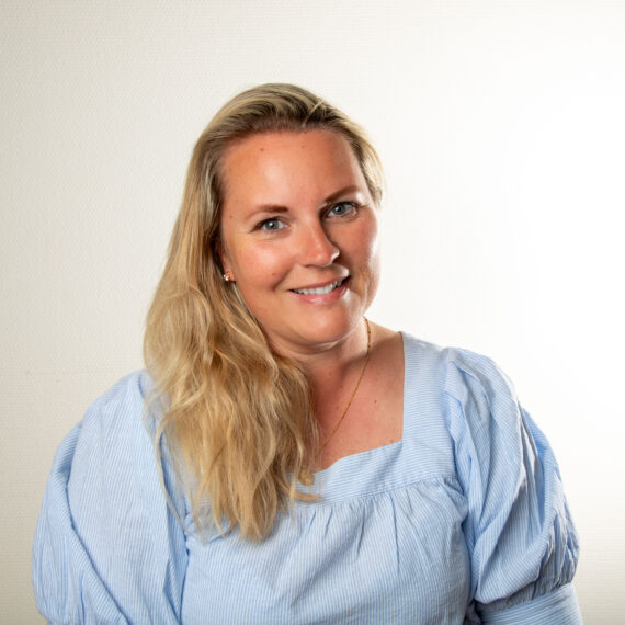 En smilende kvinne med langt blondt hår iført en lyseblå bluse med puffermer poserer foran en enkel hvit bakgrunn.