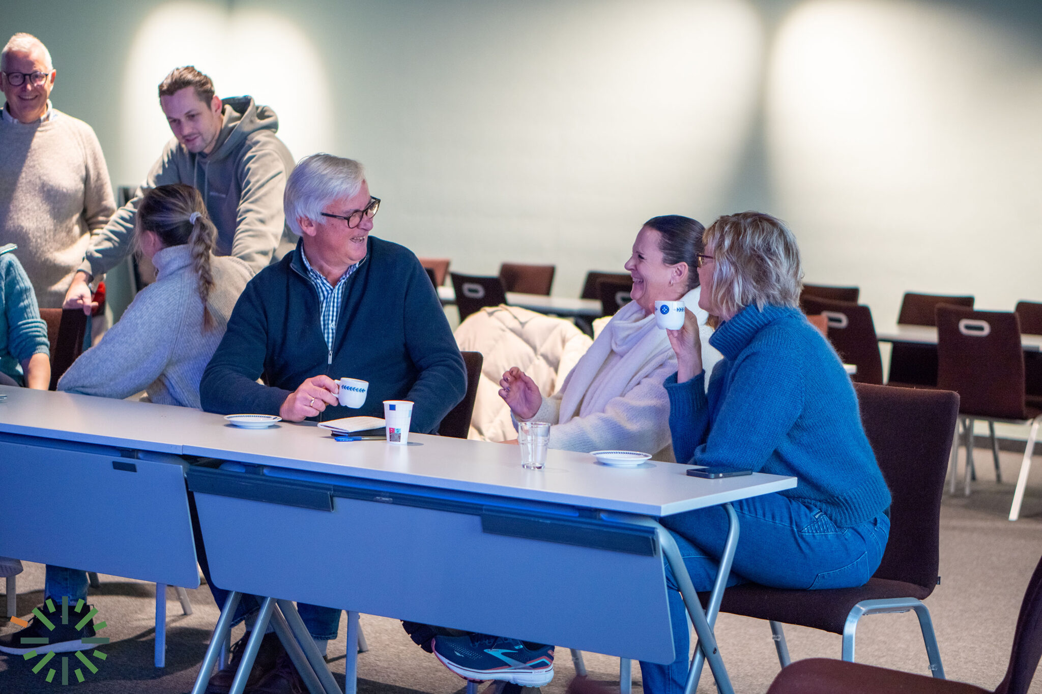En gruppe mennesker sitter og prater ved et bord i et konferanserom for kontorutleie. Tre i forgrunnen smiler med kaffekopper, mens andre samhandler i bakgrunnen. Tomme stoler er synlige bak dem.