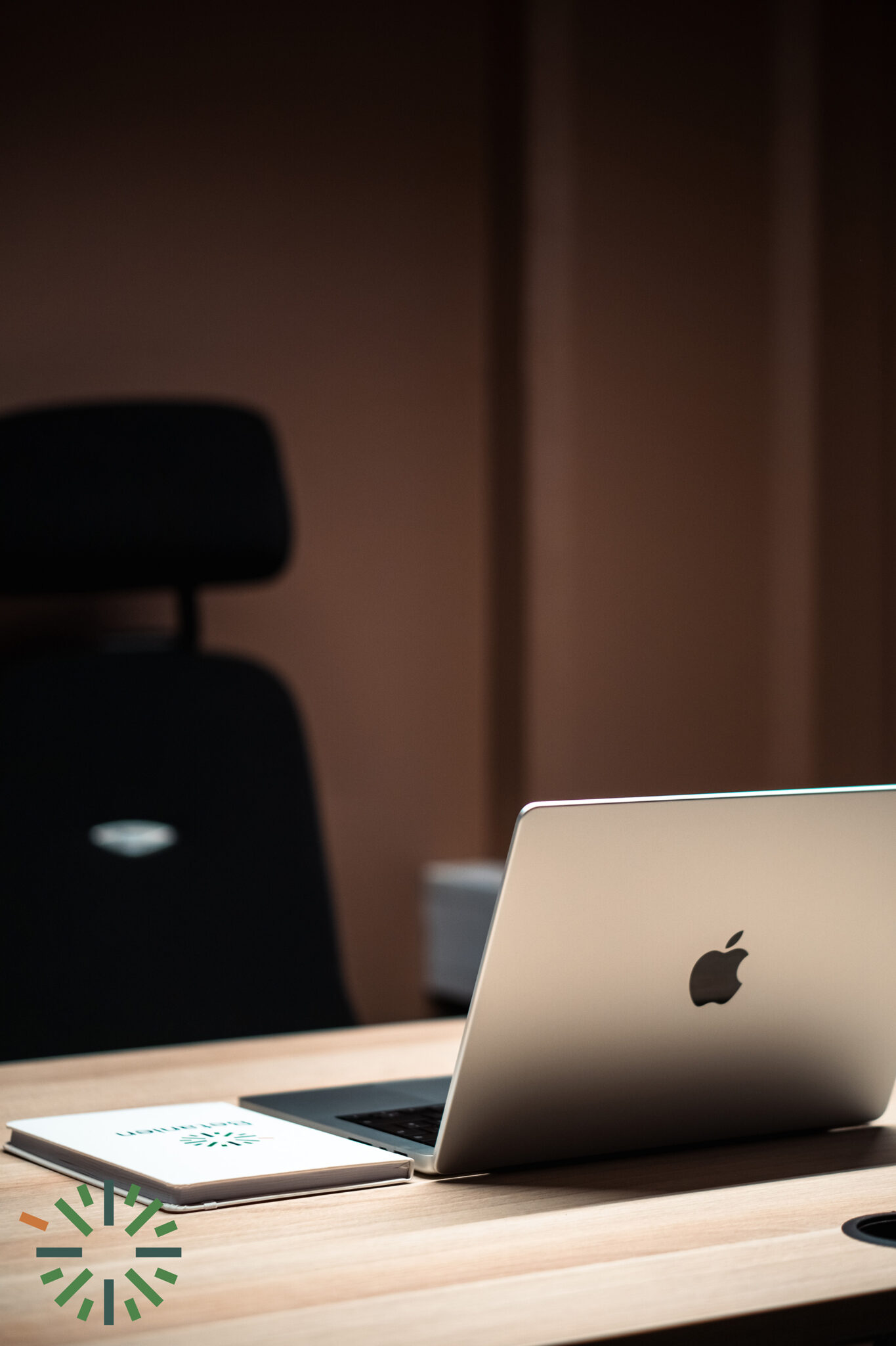 En sølvfarget MacBook står på et trebord ved siden av en lukket notatbok. I bakgrunnen fullfører en svart kontorstol den minimalistiske arbeidsplassen, perfekt for produktivitet i et moderne kontormiljø.