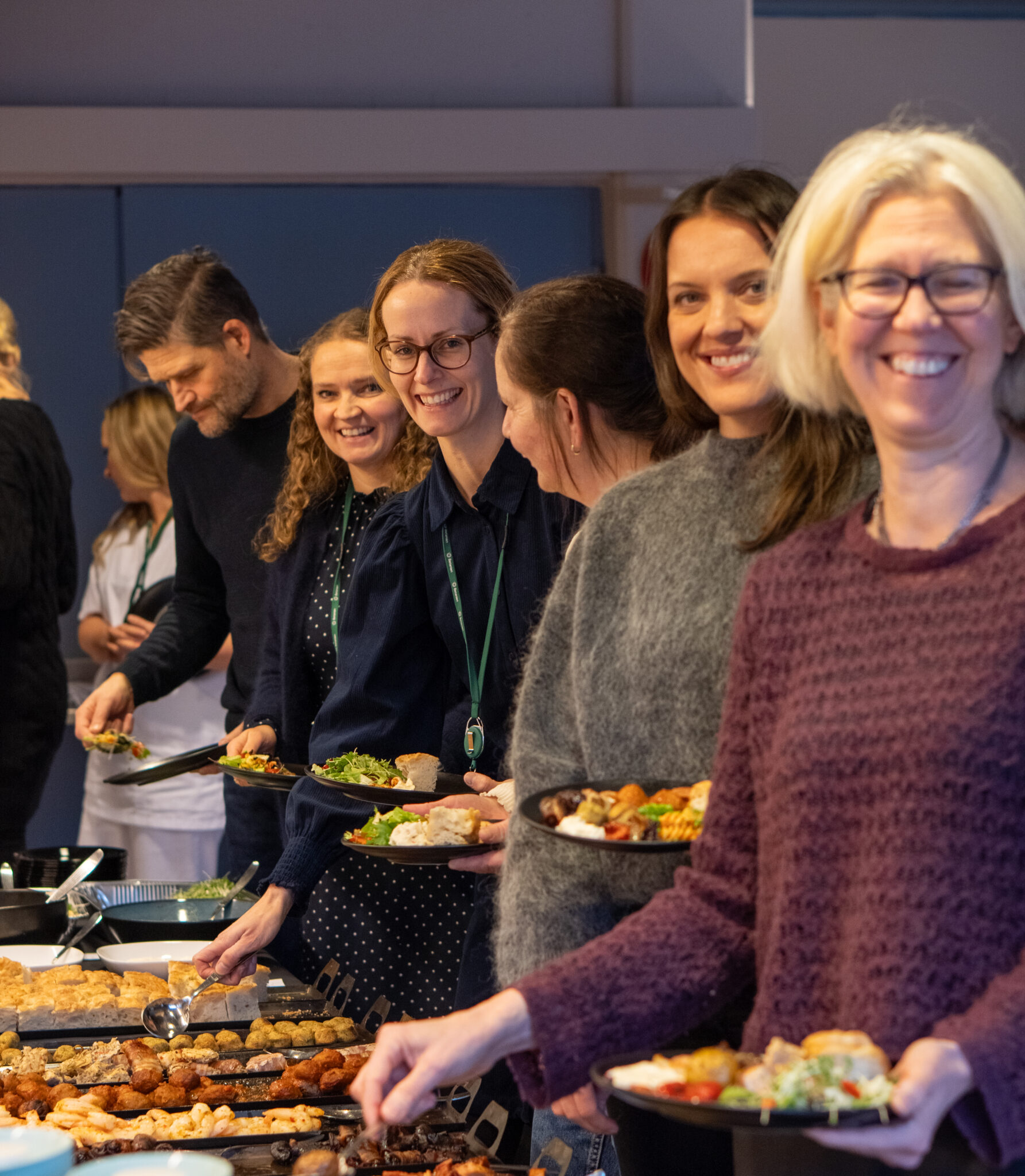 En gruppe smilende voksne står i kø ved en buffet i et kontorutleie-lokale med tallerkener fylt med diverse matvarer. Atmosfæren virker vennlig og sosial.