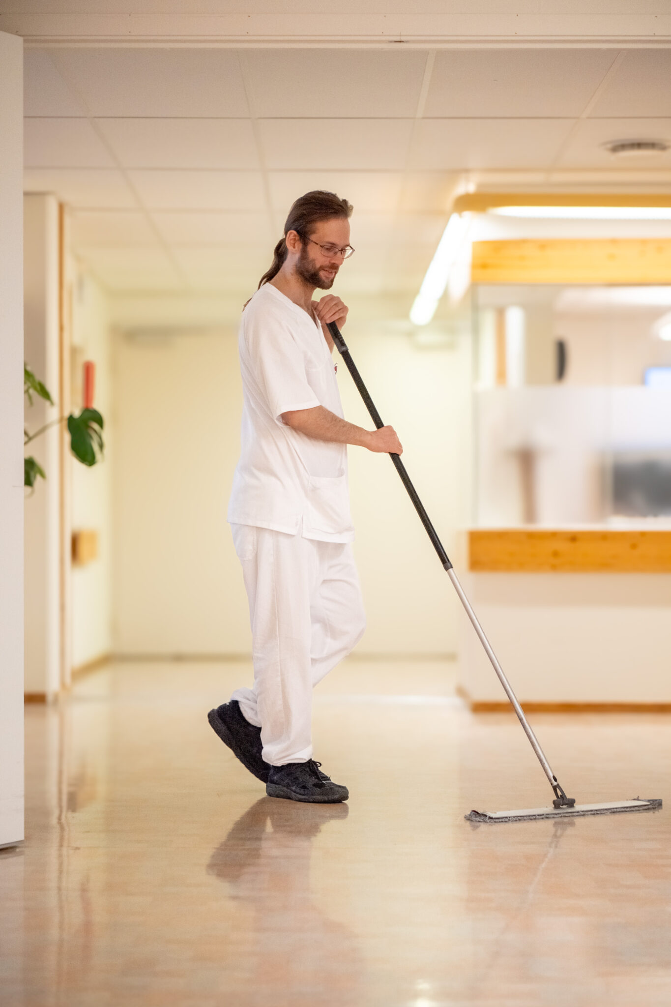 En mann i hvite skrubber og svarte sko vasker et skinnende gulv i gangen, smilende mens han ser ned. Bakgrunnen viser en godt opplyst, ren korridor med lyse vegger og en glassvegg.