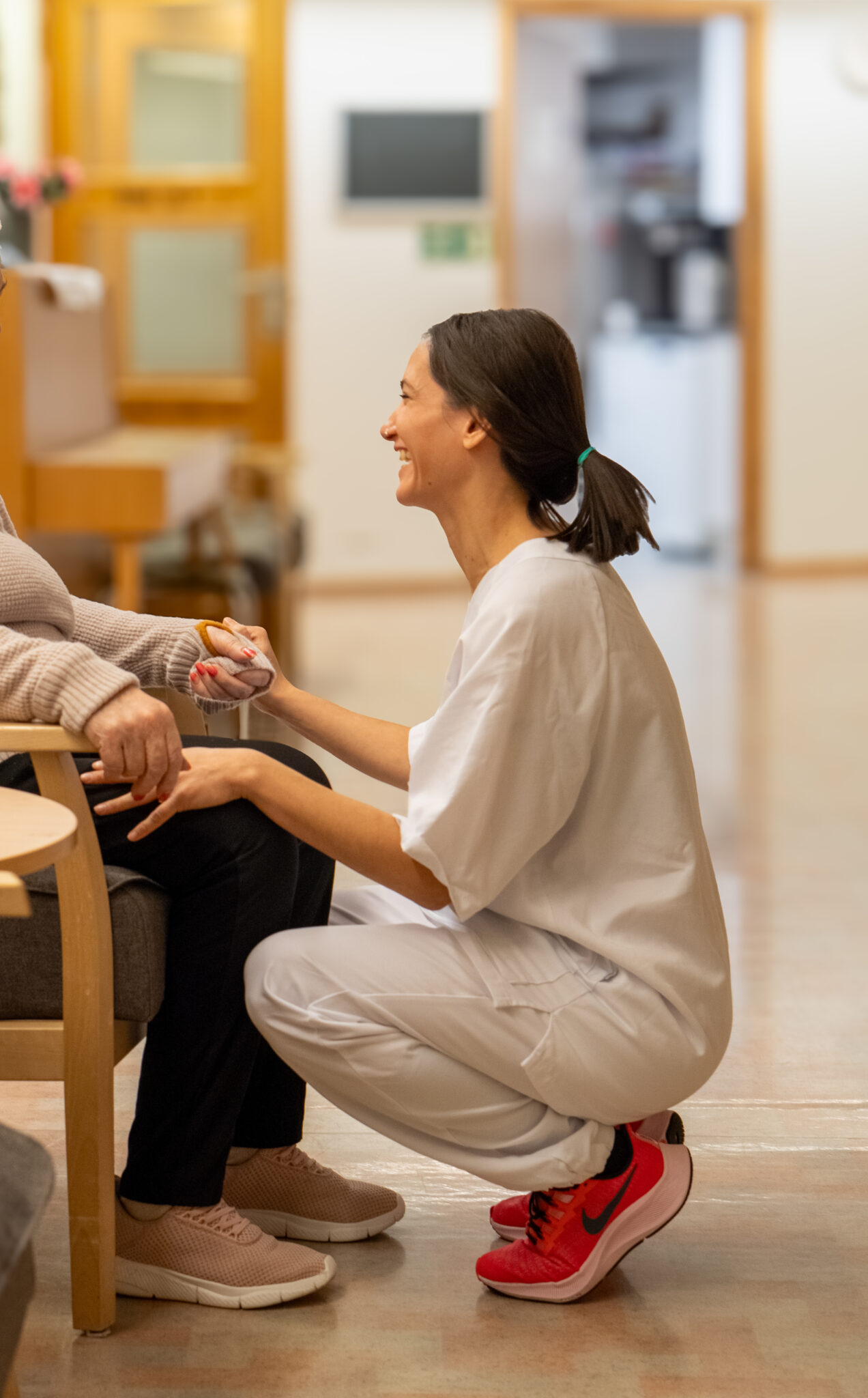 En smilende helsearbeider i hvite skrubber kneler ved siden av en eldre person som sitter i en stol og holder forsiktig hånden deres i en godt opplyst gang.