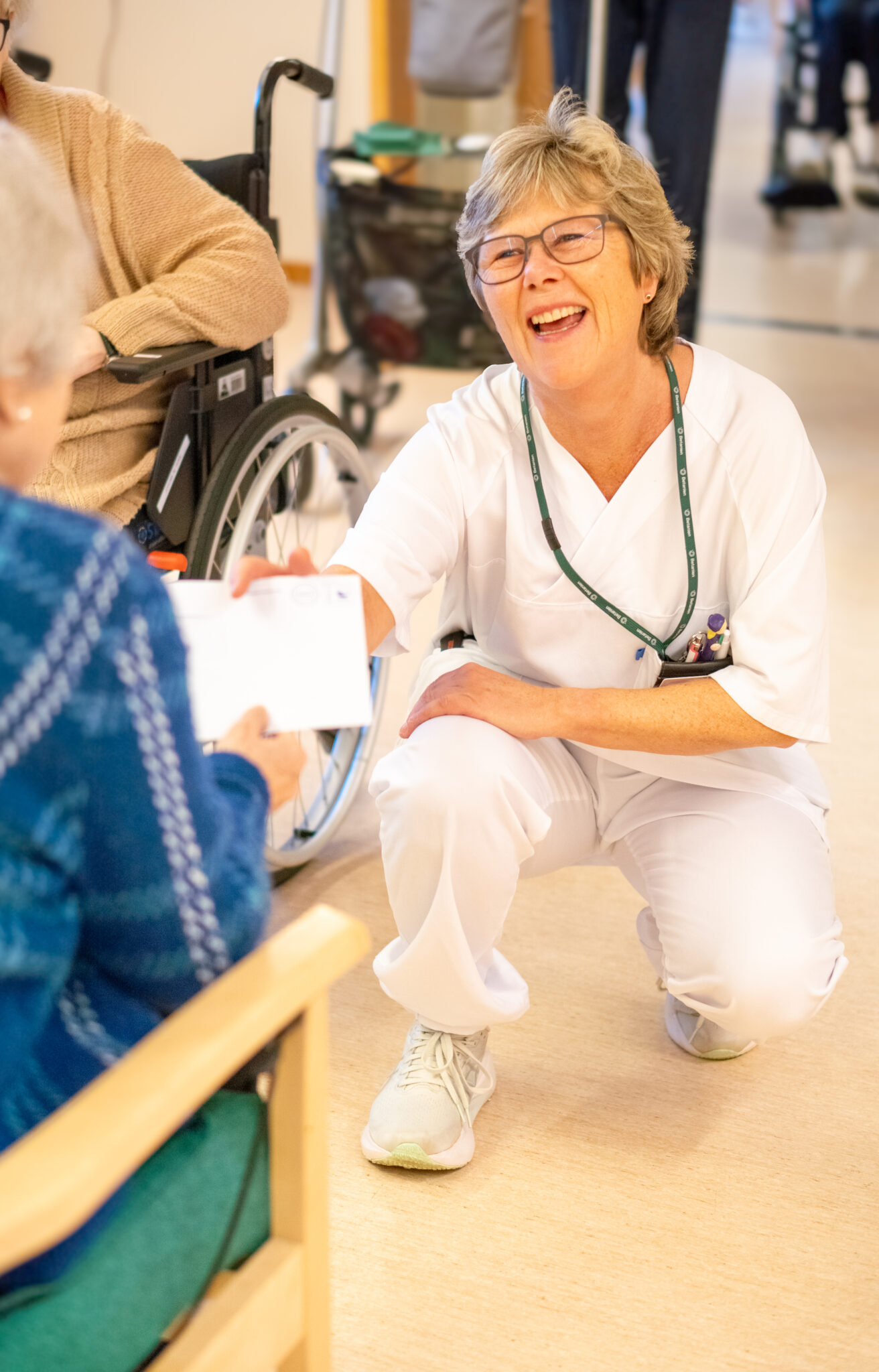 En smilende helsearbeider i hvite skrubber sitter på huk og gir et dokument til en eldre person som sitter i en stol, mens en annen person i rullestol er i nærheten.