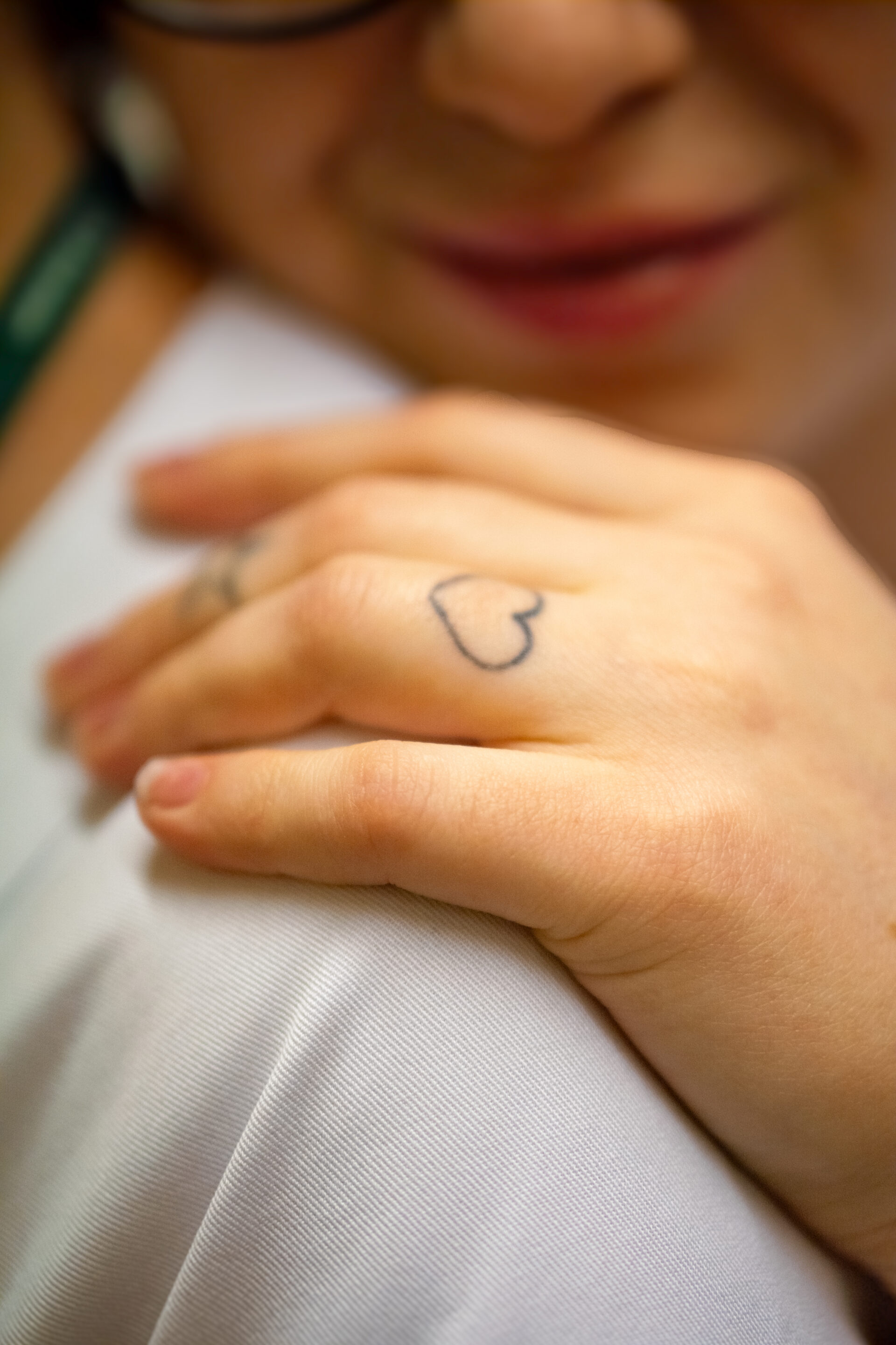 Et nærbilde av en persons hånd med en enkel hjerteformet tatovering på fingeren, som forsiktig hviler på noens skulder. Personens ansikt er delvis synlig, med et mykt smil.