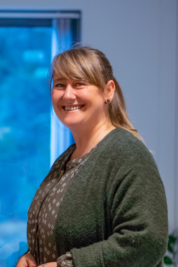 Annelen Cesilie Starefoss, med langt blondt hår og pannelugg, smiler mens hun står innendørs i en mørkegrønn genser over en mønstret bluse. Et vindu med blått lys og en del av en plante er synlige i bakgrunnen.