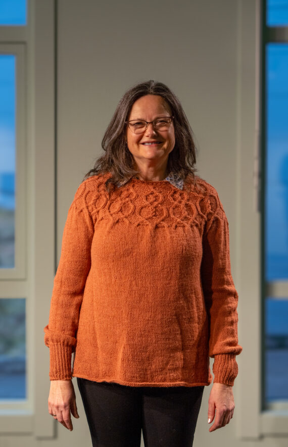 Anne Mette Borg Almeland, en kvinne med langt mørkt hår og briller, smiler mens hun står innendørs. Hun har på seg en oransje strikkegenser og svarte bukser, med store vinduer og mykt naturlig lys i bakgrunnen.