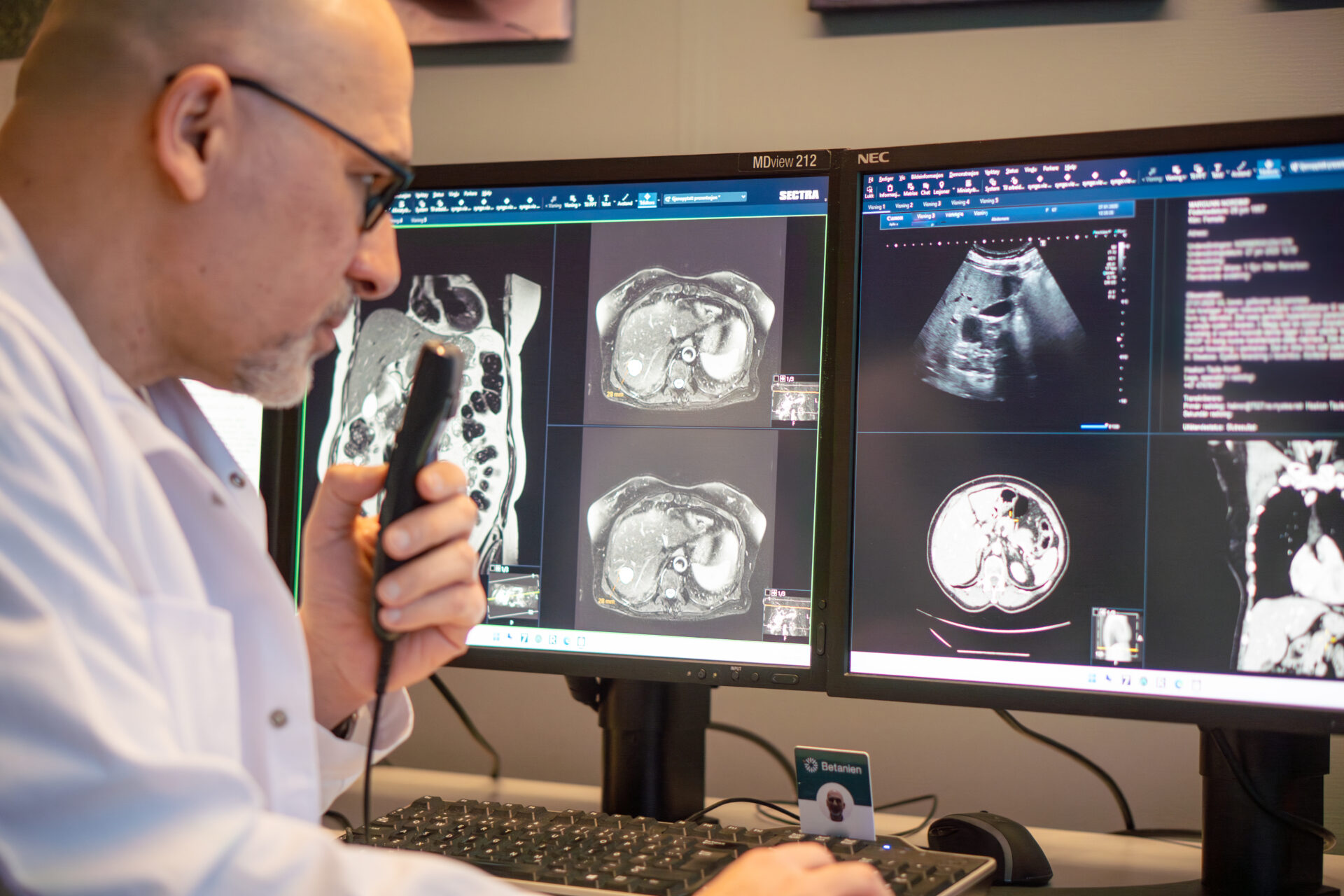 En lege i hvit frakk gjennomgår MR- og ultralydskanninger som vises på to dataskjermer, mens han holder en mikrofon for å ta opp notater eller stille en diagnose.