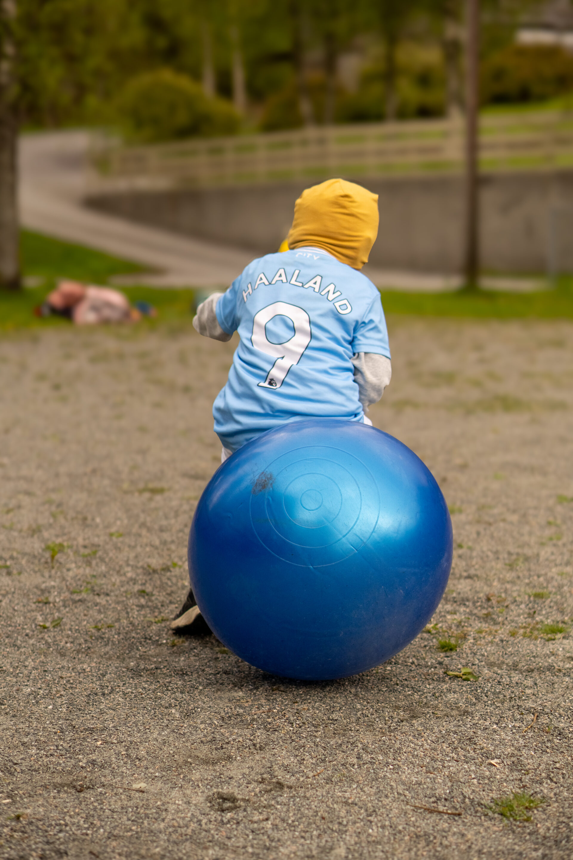 Et barn iført gul hette og en Haaland 9-fotballdrakt sitter på en stor blå treningsball utendørs på en grussti, med gress og uskarpe mennesker i bakgrunnen.