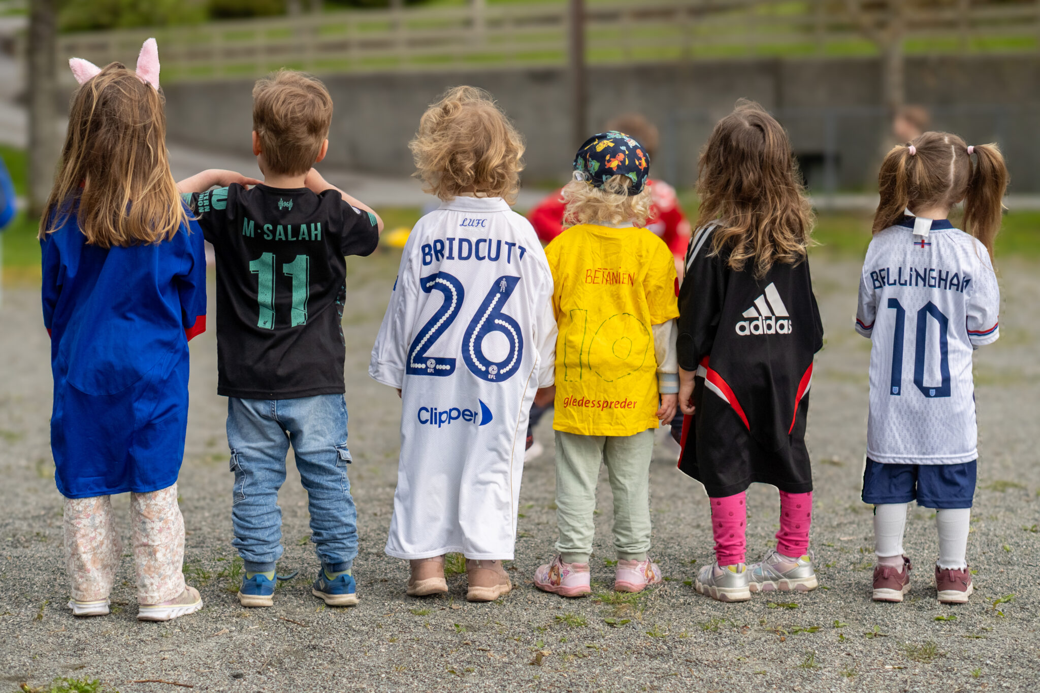Seks små barn står utendørs med ryggen mot kameraet, alle med forskjellige fotballdrakter på. De står på en grusflate og holder hender, og bakgrunnen ser ut til å være en park eller lekeplass.