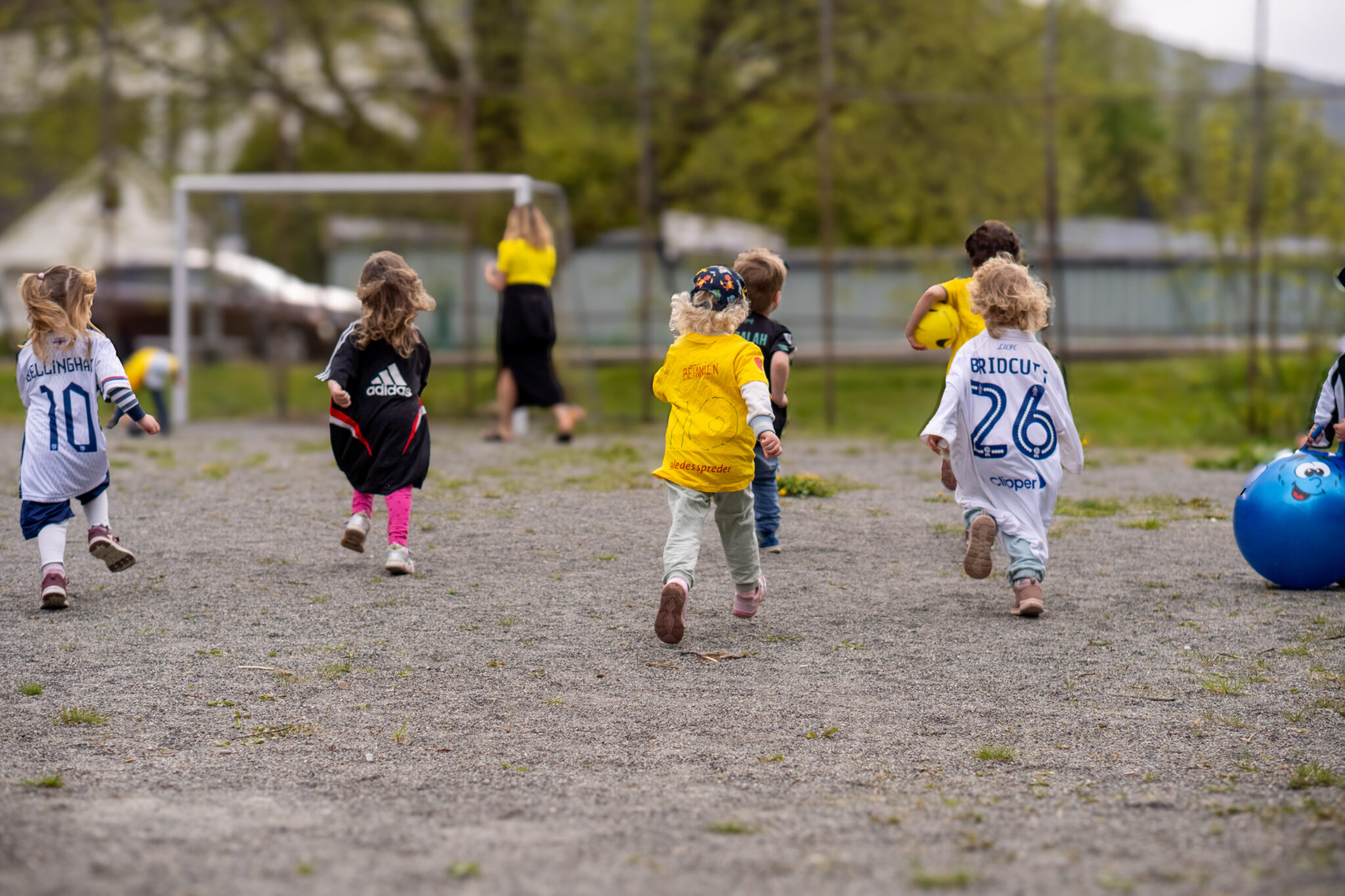 En gruppe små barn i sportsdrakter løper mot et fotballmål på en grusbane, med to voksne som står i nærheten av målet og en stor blå ball i forgrunnen. Bakgrunnen viser trær og en bygning.