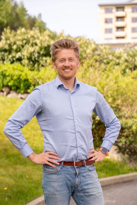 En mann med blondt hår, iført en lyseblå skjorte med knapper og jeans, står utendørs med hendene i siden og smiler til kameraet. Det er grøntområder og en bygning i bakgrunnen.