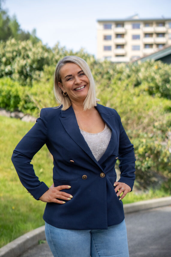 En kvinne med blondt hår står utendørs og smiler med hendene i siden. Hun har på seg en marineblå blazer over en lys topp og blå jeans. Det er grønne busker og en bygning i bakgrunnen.