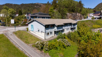 En toetasjes lyseblå bygning med skråtak og parabolantenner, omgitt av grøntområder og busker. Bygningen ligger nær en vei med flere bygninger og en skogkledd åsside i bakgrunnen.