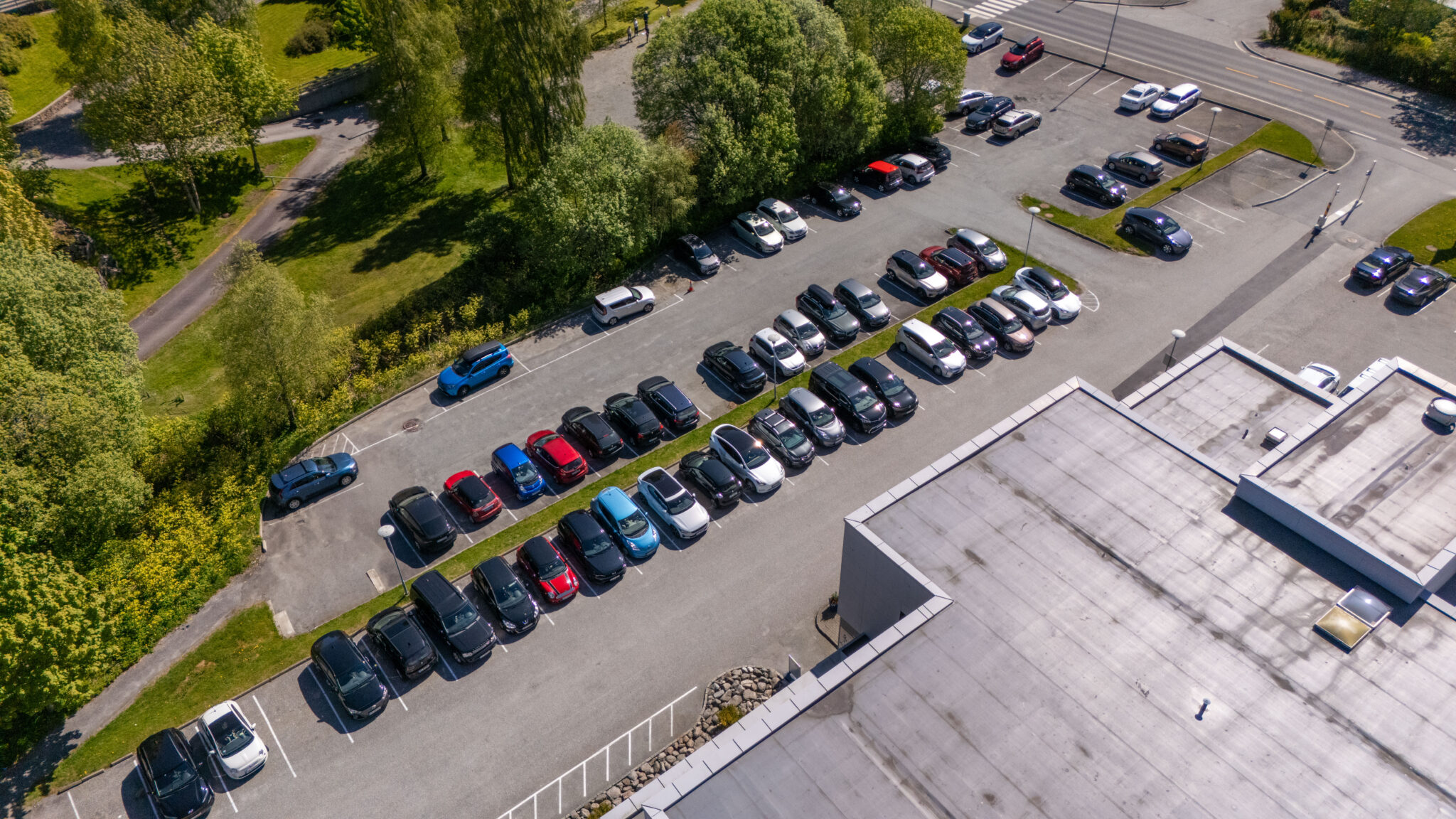 Luftfoto av en parkeringsplass fylt med parkerte biler ved siden av en stor bygning, omgitt av grøntområder og trær, med en vei og flere parkerte biler i bakgrunnen.