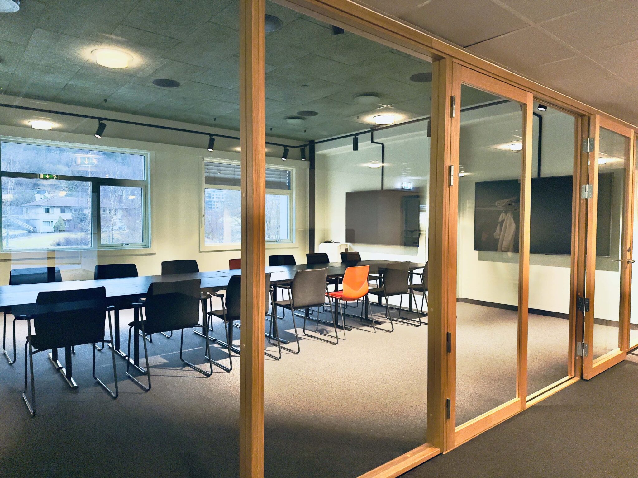 Moderne konferanserom med et stort bord omgitt av svarte stoler, hvorav én oransje stol skiller seg ut. Glassvegger omslutter rommet; store vinduer slipper inn naturlig lys og gir utsikt utover. Rommet er ryddig og tomt.