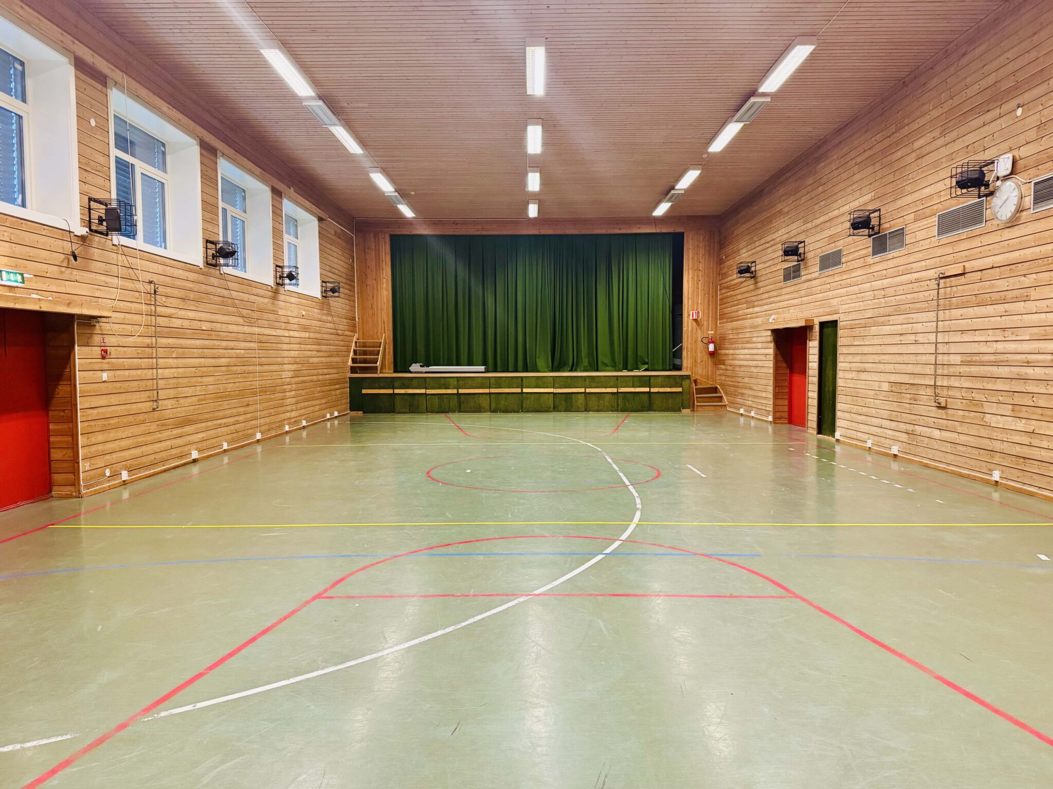 En romslig innendørs gymsal med grønt gulv merket for sport, trevegger, høye vinduer og lysrør. Ytterst i enden er det en hevet scene med et lukket grønt forheng.