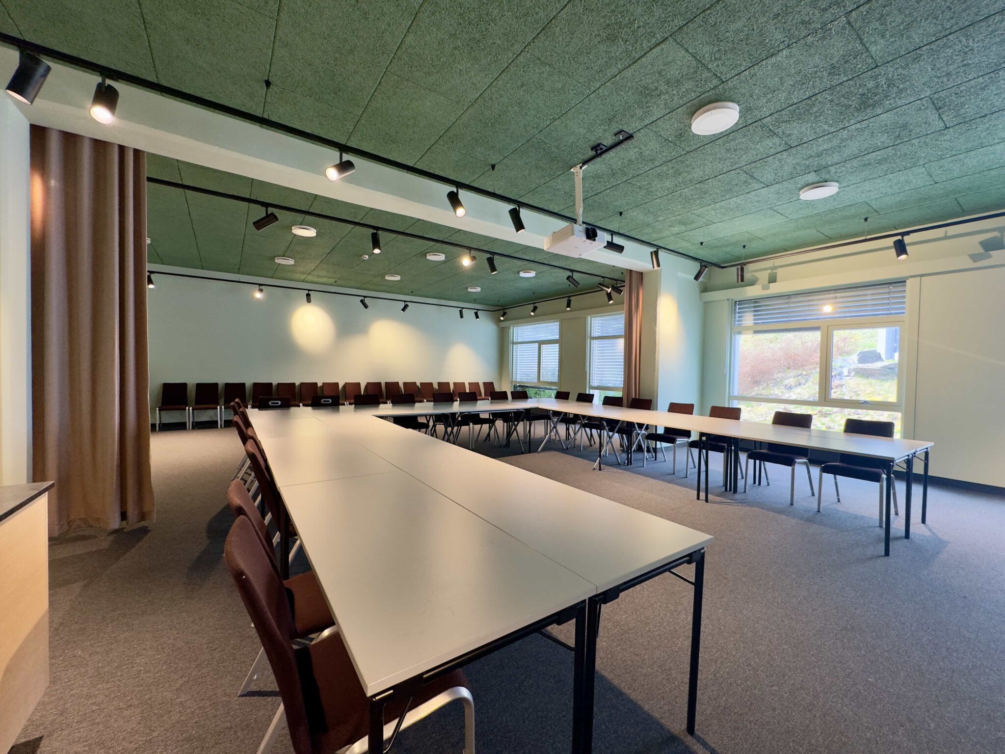 Et moderne konferanserom med U-formede bord og brune stoler, skinnebelysning i taket, store vinduer som slipper inn naturlig lys, grått teppe og grønt aksenttak.