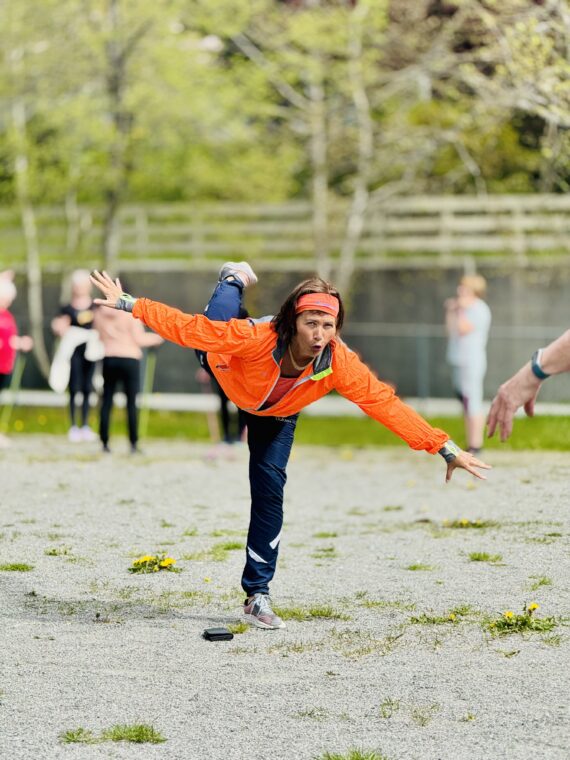 En person i oransje jakke og marineblå bukser balanserer på ett ben utendørs og strekker ut armene for å holde balansen. Det er flere personer som er uskarpe i bakgrunnen på en grusoverflate med grøntområder.