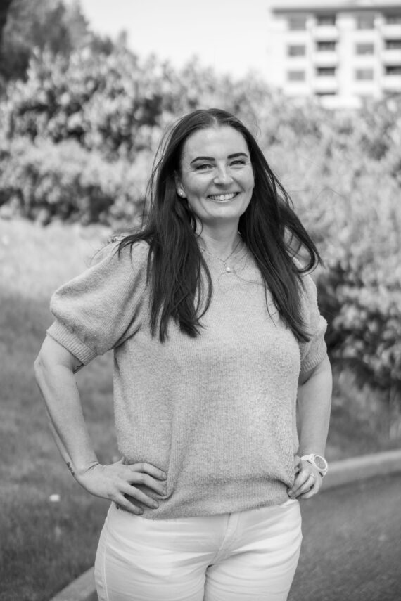 Vivian Hestholm, en kvinne med langt mørkt hår, smiler utendørs med hendene på hoftene. Hun har på seg en kortermet genser og lyse bukser. Bakgrunnen i dette svart-hvitt-bildet viser trær og en uskarp bygning.
