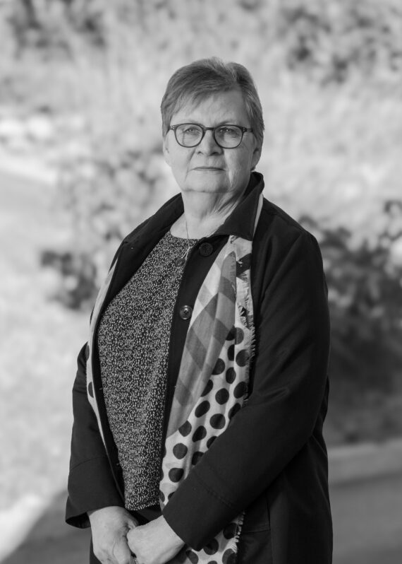Svart-hvitt-bilde av en eldre kvinne med briller, et mønstret skjerf, en mørk frakk og en mønstret topp, stående utendørs med uskarpe trær og løvverk i bakgrunnen.