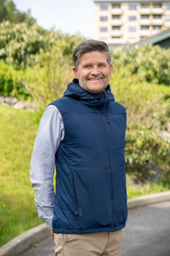 En mann med kort grått hår og skjegg smiler mens han står utendørs. Han har på seg en blå vest over en lysegrå skjorte og beige bukser, med grøntområder og en bygning i bakgrunnen.