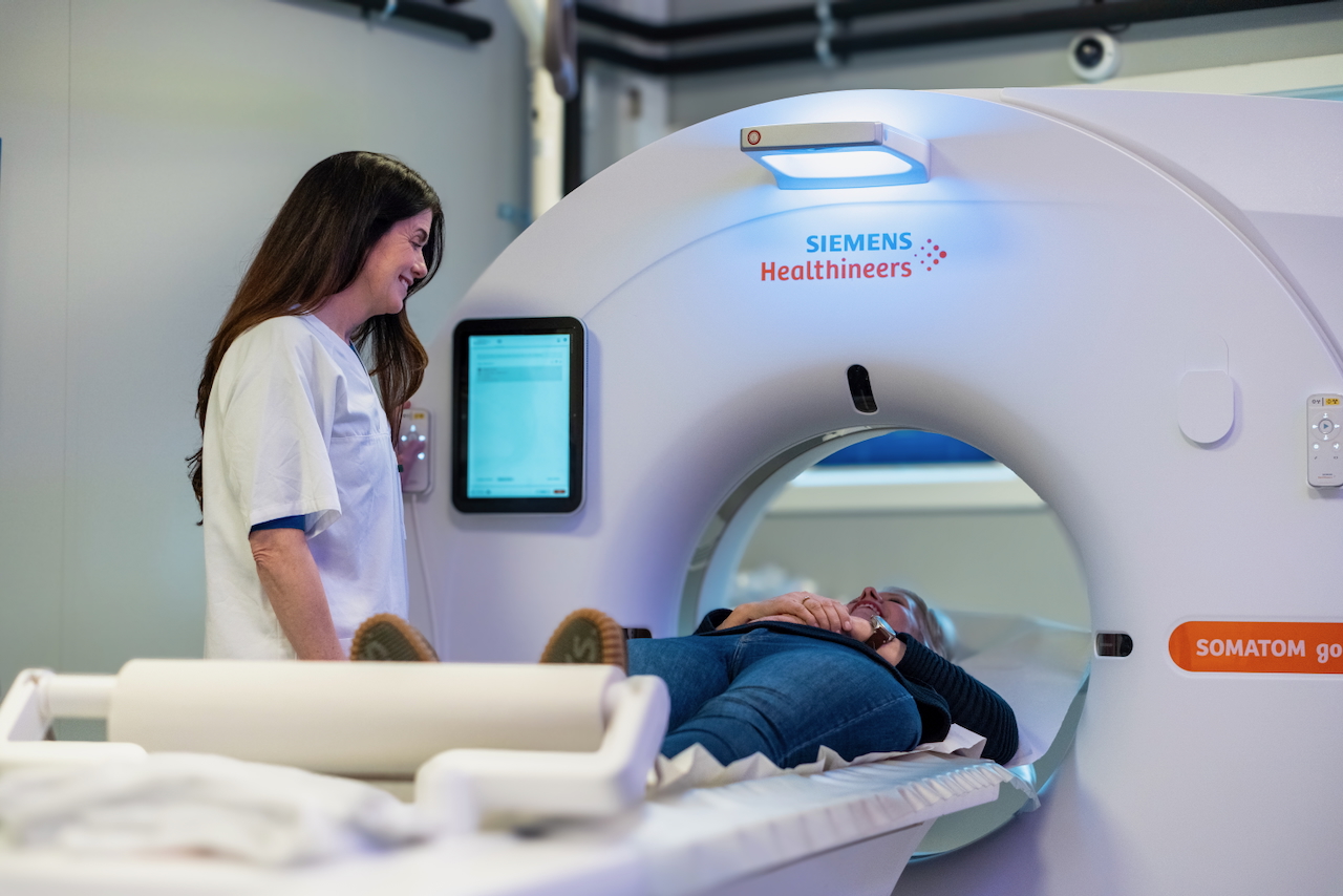 En helsepersonell fra Stiftelsen Betanien står ved siden av en pasient som ligger på et skyvebord og forbereder seg til en skanning inne i en Siemens Healthineers SOMATOM medisinsk bildebehandlingsmaskin. Pasienten ser avslappet ut og smiler.