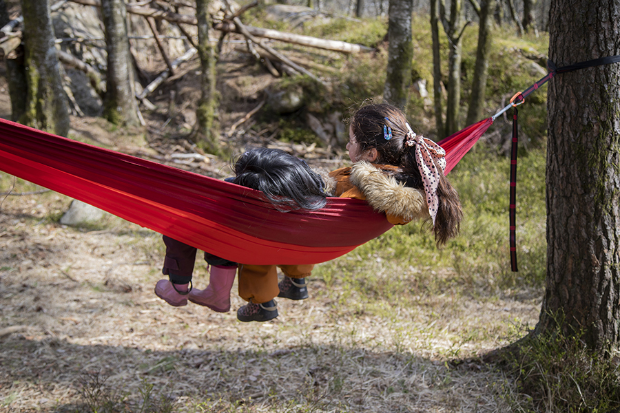 To barn ligger sammen i en knallrød hengekøye bundet mellom trær i en skog. De har på seg jakker og støvler, og det ene barnets hår er bundet opp med et skjerf, slapper av og ser bort fra kameraet.