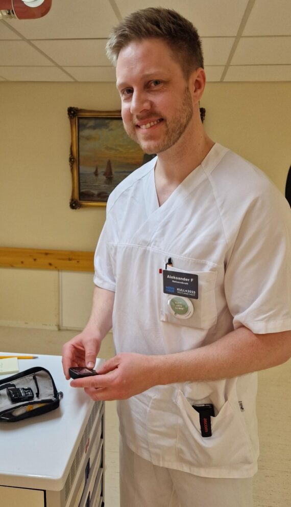 En smilende helsearbeider, Aleksander Fløteberget, i hvite skrubber står i en klinisk setting med et medisinsk utstyr i hånden. Et navneskilt er synlig på brystet hans, og et maleri pryder veggen bak ham.
