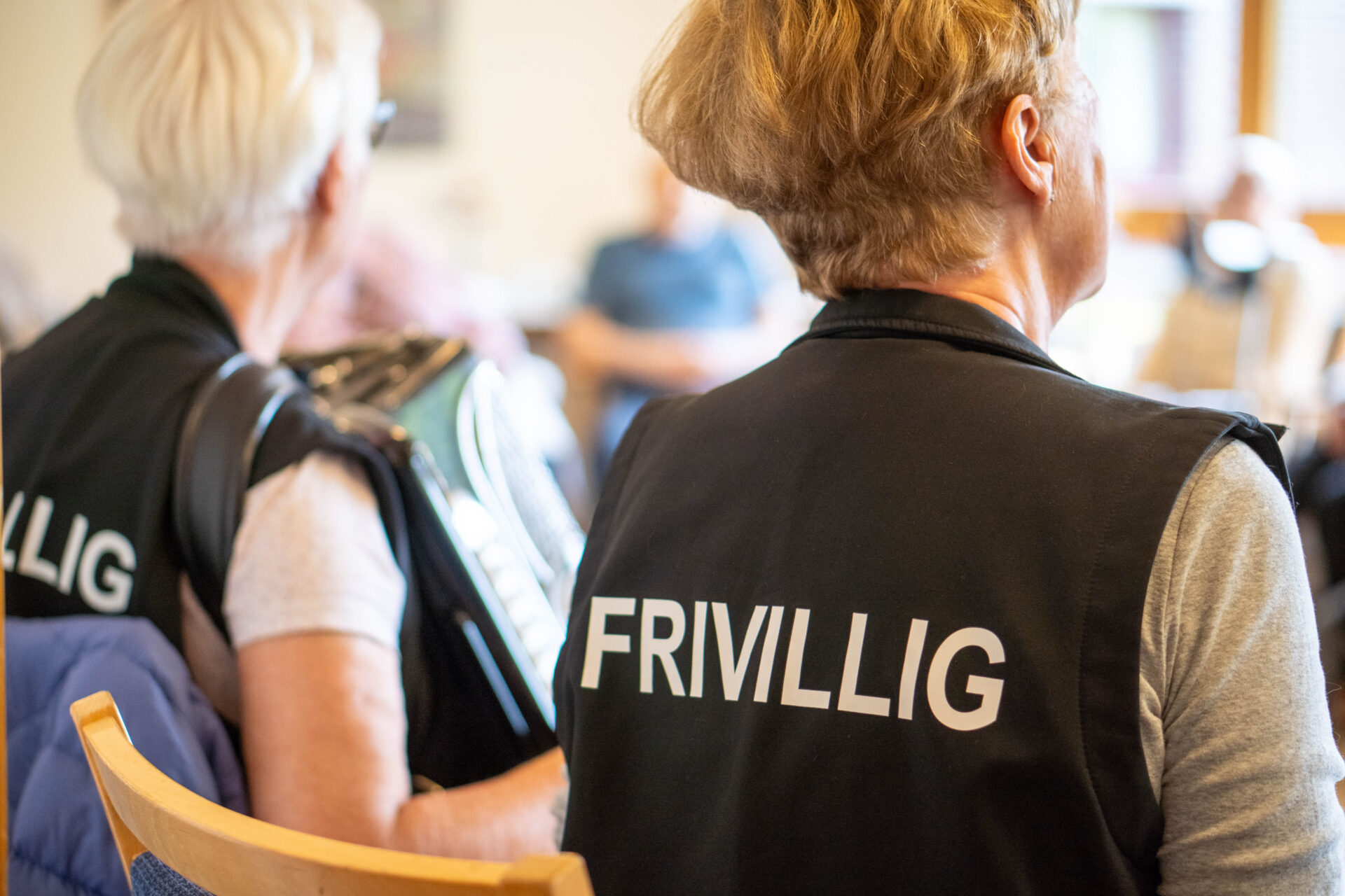 To frivillige kvinner sitter med ryggen til kameraet i et lyst rom, iført vester med teksten “FRIVILLIG” trykket på ryggen. Den ene holder et trekkspill. I bakgrunnen skimtes eldre personer i samtale og aktivitet, noe som tyder på en sosial samling, trolig på et sykehjem eller aktivitetssenter.