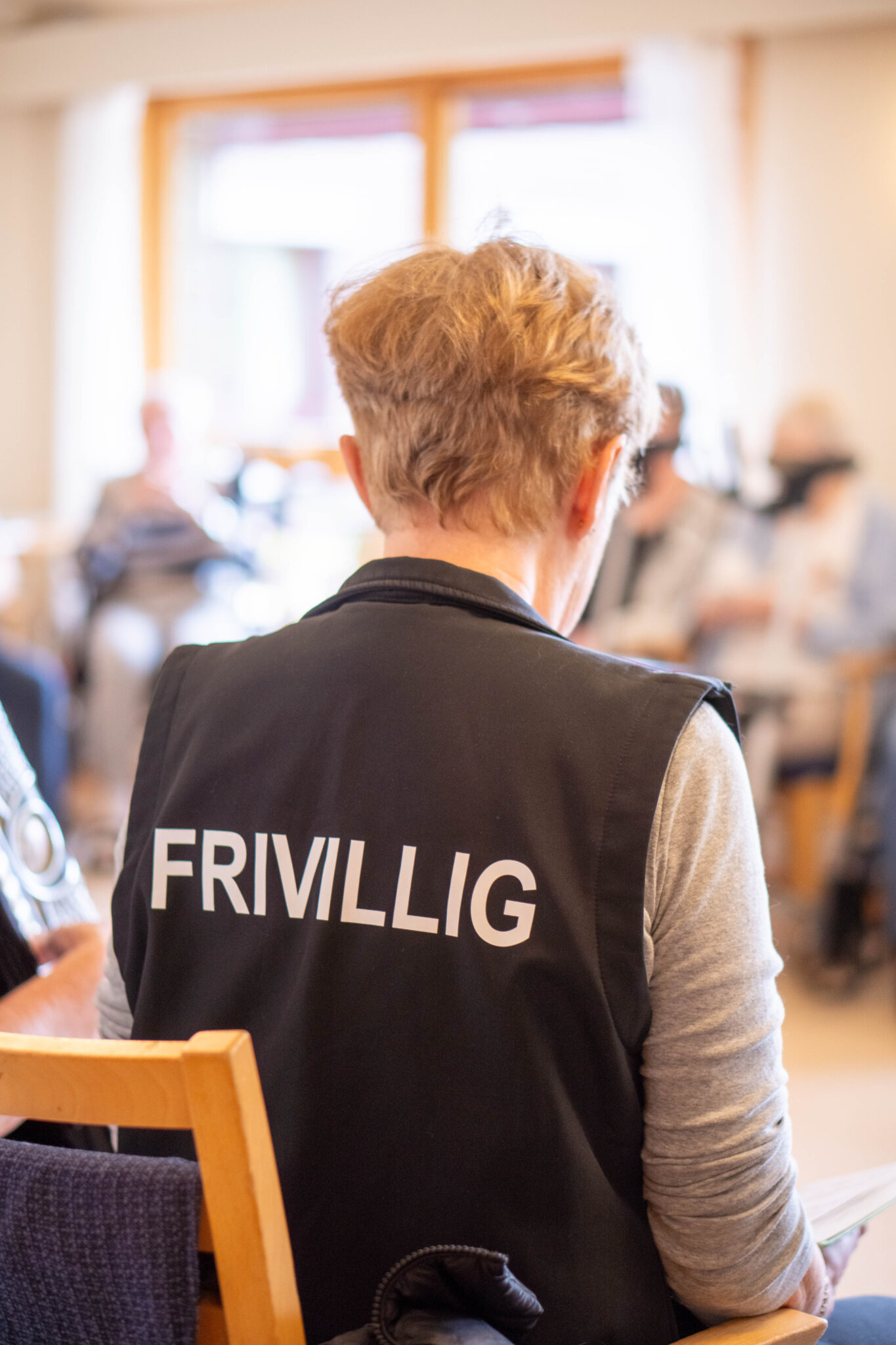 To frivillige kvinner sitter med ryggen til kameraet i et lyst rom, iført vester med teksten “FRIVILLIG” trykket på ryggen. Den ene holder et trekkspill. I bakgrunnen skimtes eldre personer i samtale og aktivitet, noe som tyder på en sosial samling, trolig på et sykehjem eller aktivitetssenter.