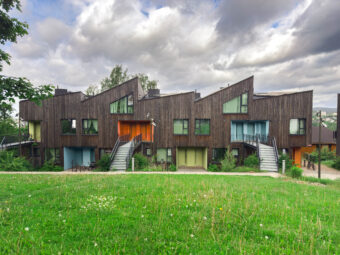 Moderne rekkehus med trekledning og skrå tak, sett fra en grønn plen. Hver bolig har fargerike inngangsdører i blant annet oransje, grønn og blå, som gir et livlig uttrykk. Store vindusflater gir godt med lys inn i boligene, og trappeoppganger leder opp til andre etasje. Himmelen er delvis skyet, og bak bygningene sees skog og et utsyn over en by eller dal.