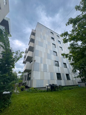 En moderne leilighetsbygning i flere etasjer med rutete fasade i lysegrå toner, omgitt av grønt gress og trær under en overskyet himmel. Utemøbler er synlige på plenen.
