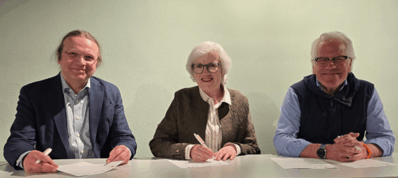 Tre voksne, to menn og en kvinne, sitter smilende ved et bord mens de signerer dokumenter. De er kledd i forretningsantrekk mot en ensfarget bakgrunn.