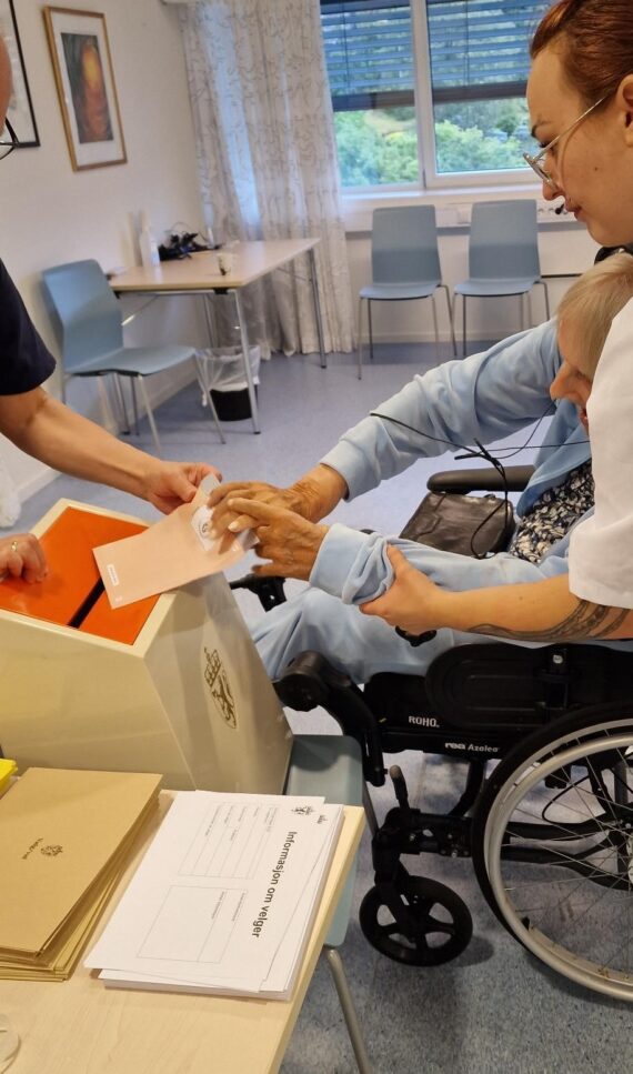 En eldre person i rullestol, assistert av en pleier, legger en stemmeseddel i en stemmeurne i et lyst rom. Dokumenter ligger på et bord i nærheten.