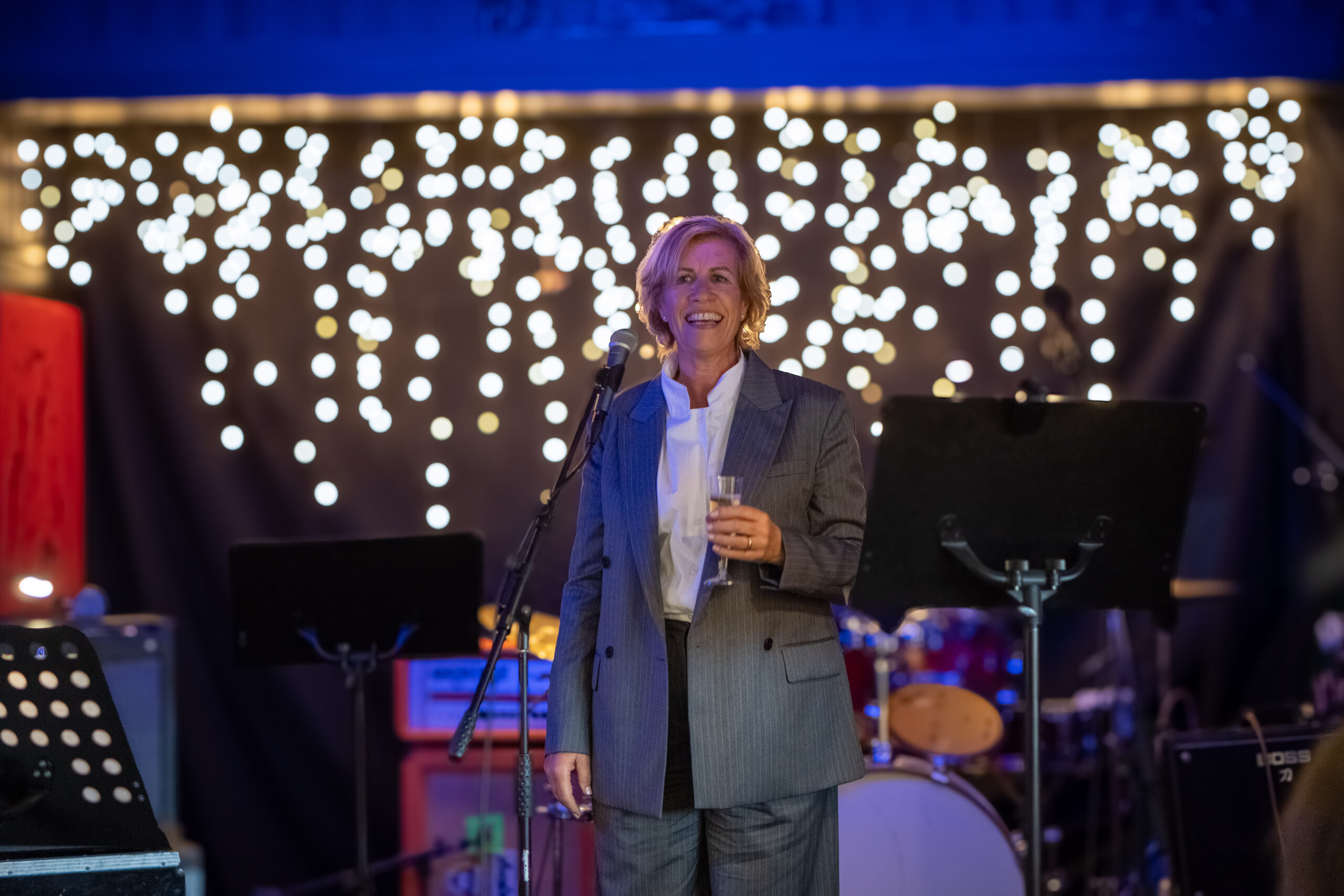 En person i grå dress står smilende på scenen med en drink i hånden, med mikrofoner og lyskastere i bakgrunnen.