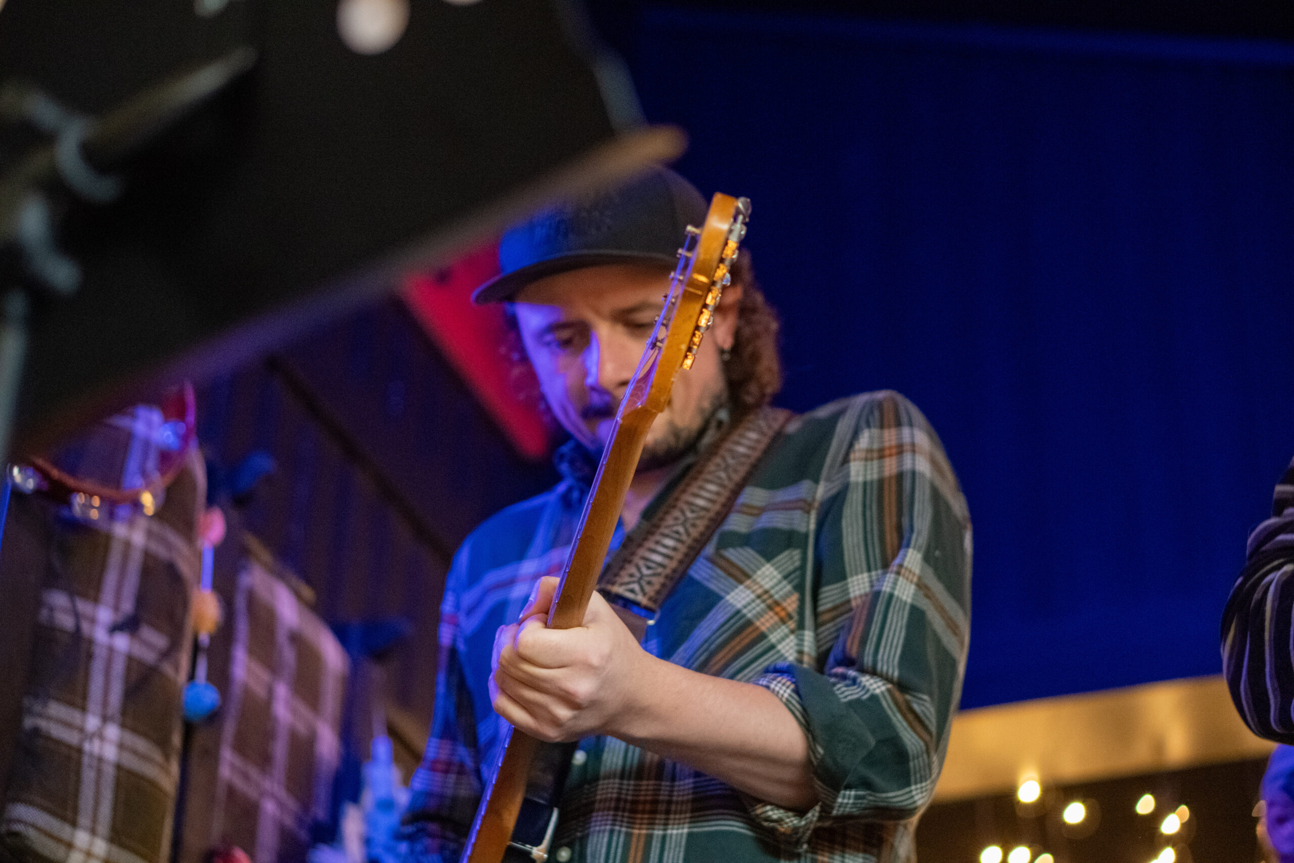 En person i rutete skjorte og caps spiller elektrisk gitar på scenen med blå belysning i bakgrunnen.