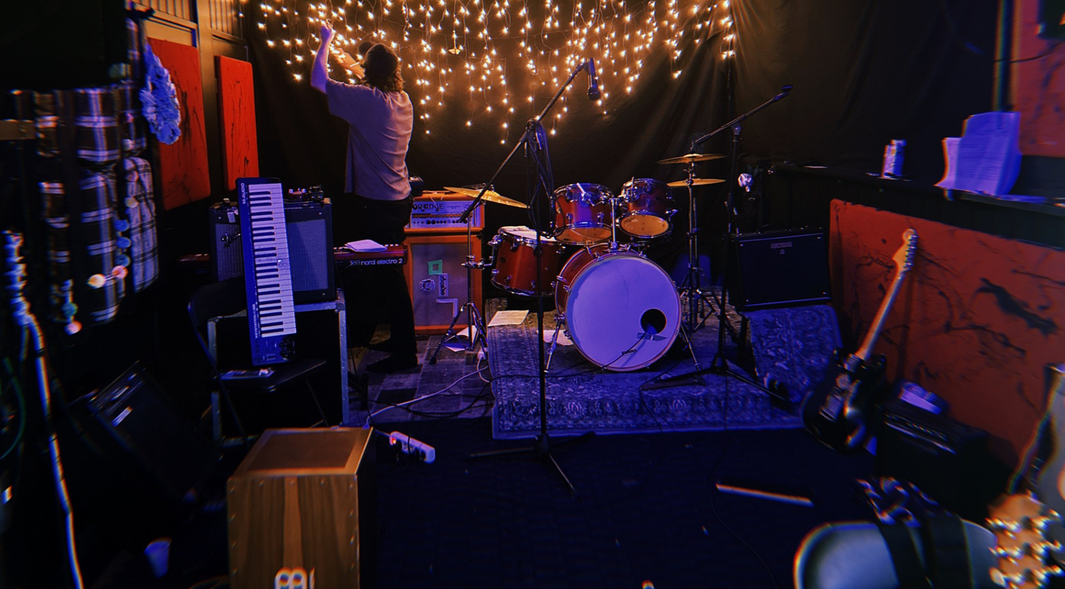 En person justerer lysstrenger over et trommesett i et svakt opplyst musikkstudio med ulike instrumenter og utstyr plassert rundt i rommet.