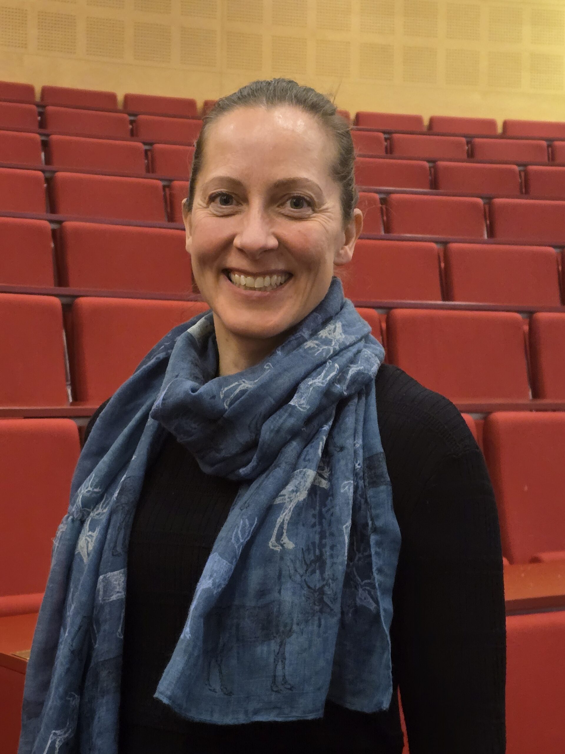 En kvinne med blått skjerf og svart topp står smilende foran røde seter i et auditorium.