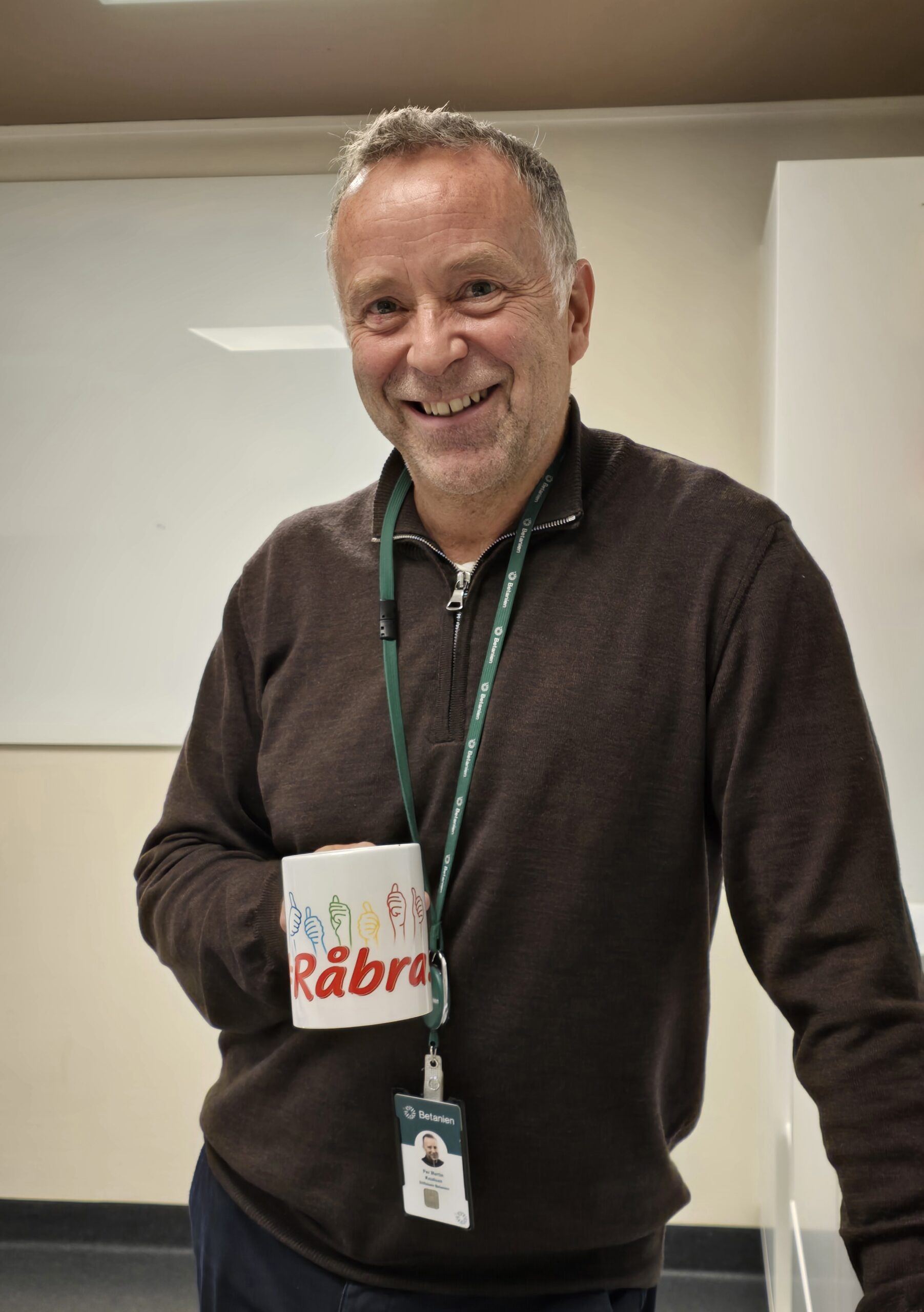 En mann i mørk genser smiler mens han holder et krus merket Råbra i en innendørssituasjon. Rundt halsen bærer han et nøkkelbånd med et ID-skilt.