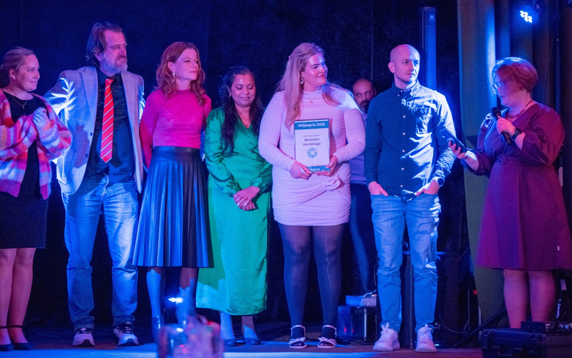 Seks personer står på en scene i blått og lilla lys; en kvinne holder et innrammet diplom som feirer Betanien barnehages miljøpris 2025, mens en annen snakker i en mikrofon.