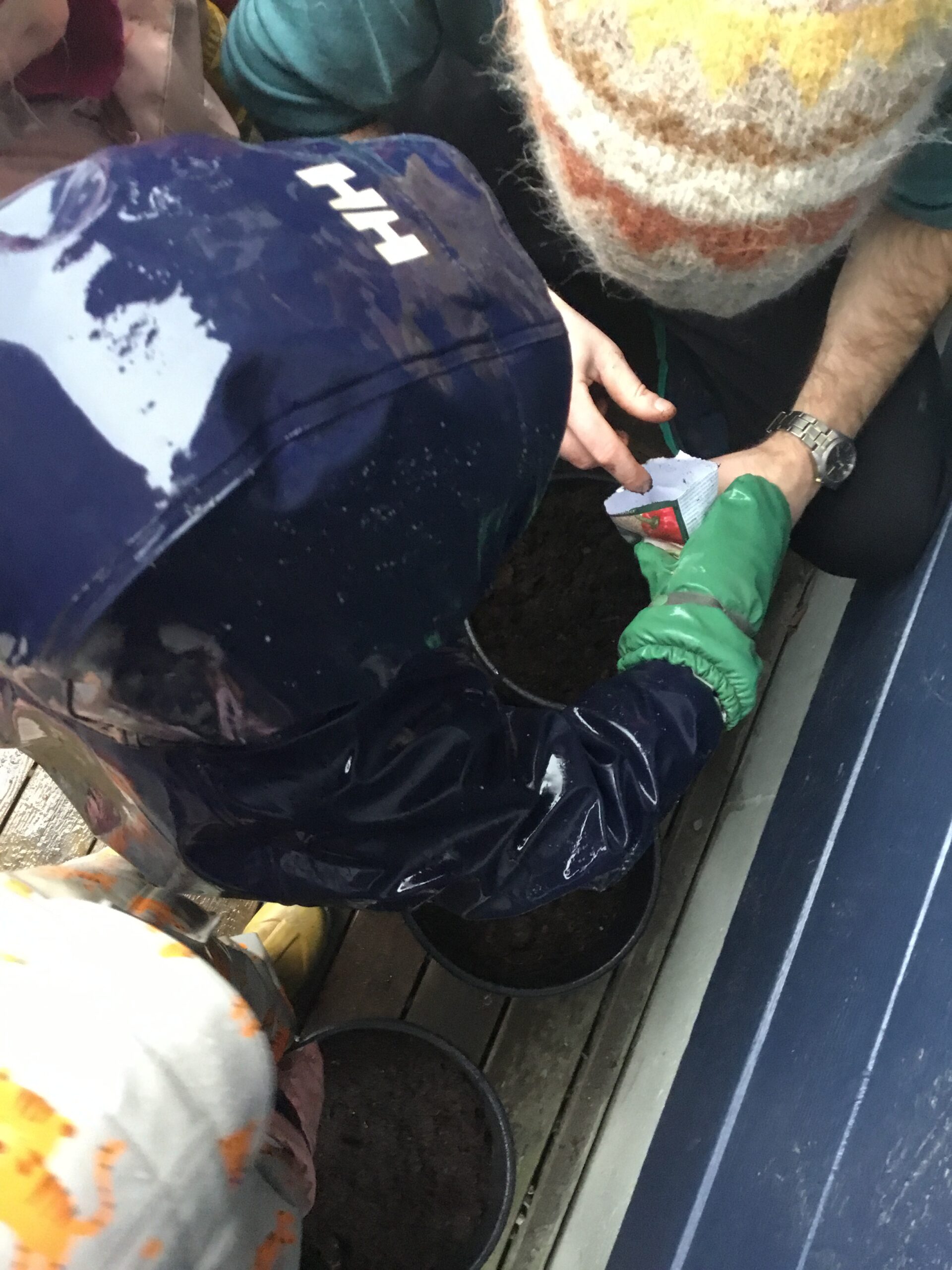 Et barn i regnfrakk og en voksen planter frø sammen i en potte; den voksne holder i en frøpakke mens barnet strekker seg mot jorden.