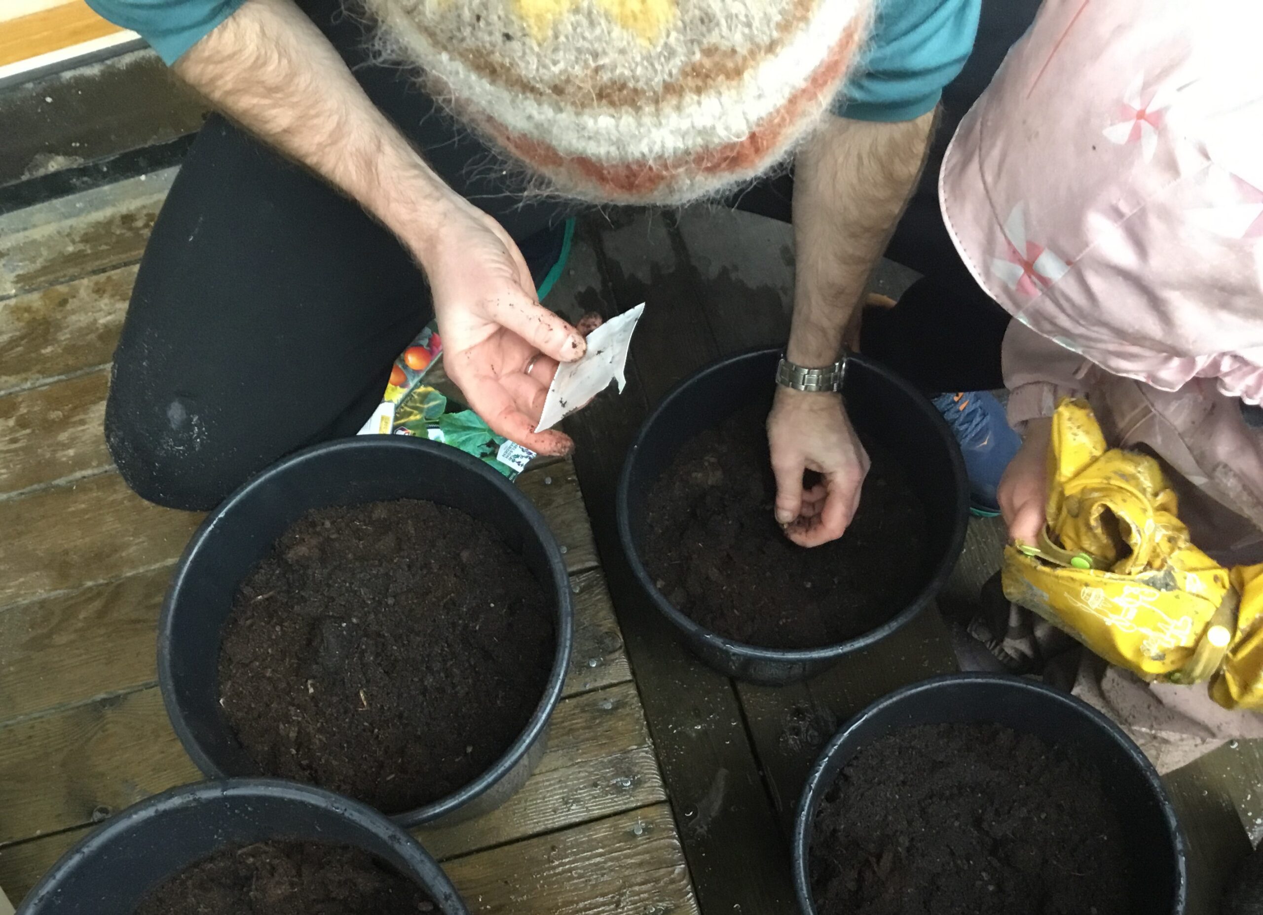 To personer, en voksen og et barn, planter frø i jordfylte potter på en treflate, mens den ene holder en frøpakke og den andre en pose med frø.