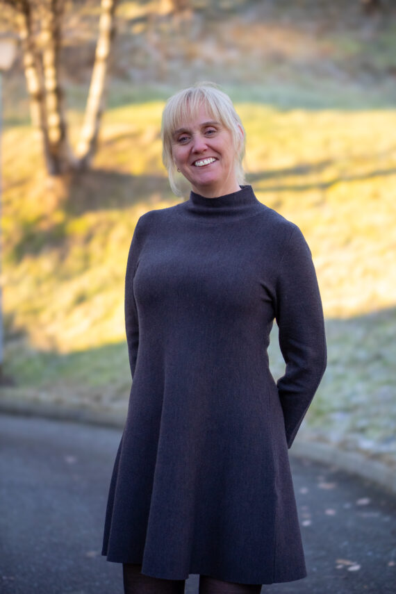 En kvinne med blondt hår, iført en langermet mørk kjole, står utendørs på en asfaltert sti og smiler til kameraet med trær og gress i bakgrunnen.