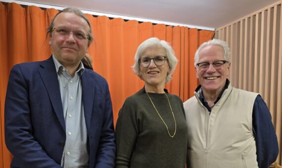 Tre voksne står innendørs foran en oransje gardin og smiler til kameraet. De to mennene bærer briller, kvinnen har kort, hvitt hår og et langt halskjede.