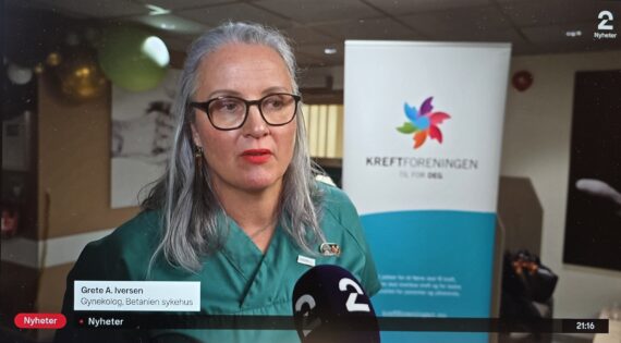 En kvinne med grått hår og briller snakker i en mikrofon. Bak henne henger et banner med en fargerik logo og teksten "Kreftforeningen". TV2 nyhetene viser hennes navn og tittel, mens hun snakker om #sjekkdeg-kampanjen.