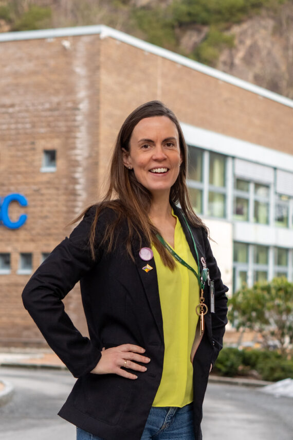 Karoline Heggøy, en kvinne med langt brunt hår, iført svart blazer og gul topp, står utenfor en murbygning og smiler til kameraet.