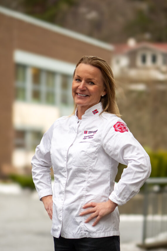 Liv Hege Olsen, kledd i hvit kokkejakke, står utendørs og smiler til kameraet, med bygninger og grøntområder mykt uskarpt i bakgrunnen.
